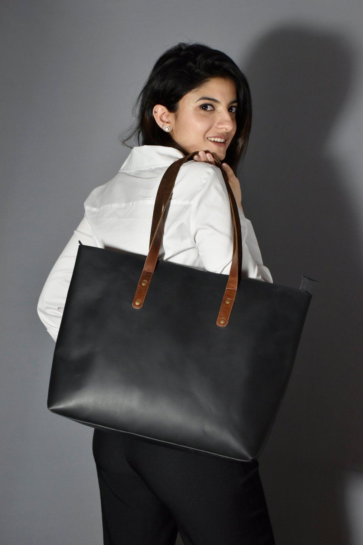 May include: A black leather tote bag with brown leather straps and metal accents. The bag is being held by a person wearing a white shirt.
