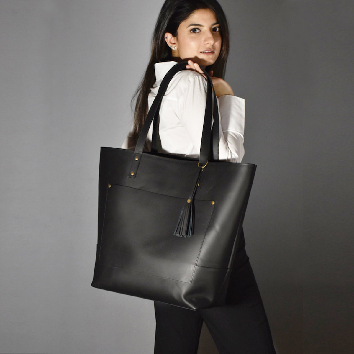 May include: A black leather tote bag with a rectangular shape and a front pocket. The bag has two long handles and a decorative tassel. Gold-colored rivets and a ring accent the design. The bag is carried over the shoulder.