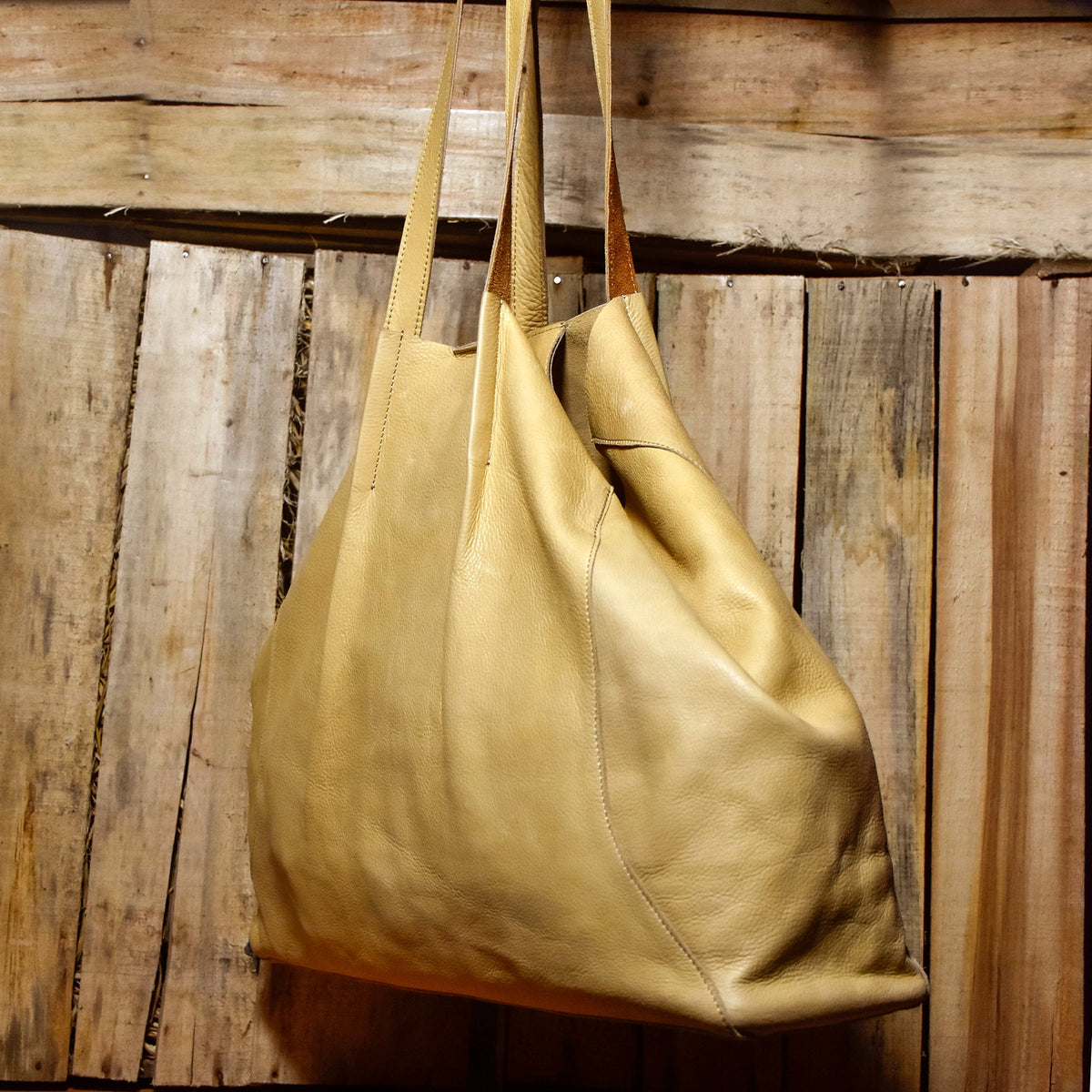 May include: A large, light tan leather tote bag with long straps, hanging against a rustic wooden background. The bag has a soft, slouchy design and visible stitching details. The leather appears to have a natural grain.