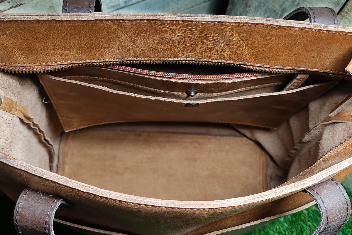 May include: A tan leather tote bag interior featuring a top zipper closure, a central pocket with a snap button, and dark brown leather handles. The bag has a smooth, unlined base and visible stitching along the edges of the leather compartments.