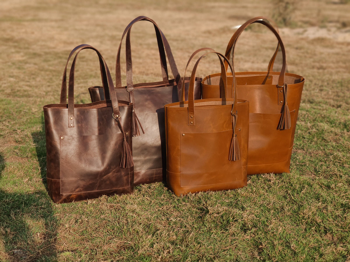 May include: Four brown leather tote bags with tassels. The bags are arranged in a row from largest to smallest. The bags are all made of the same type of leather, but they have different shades of brown.