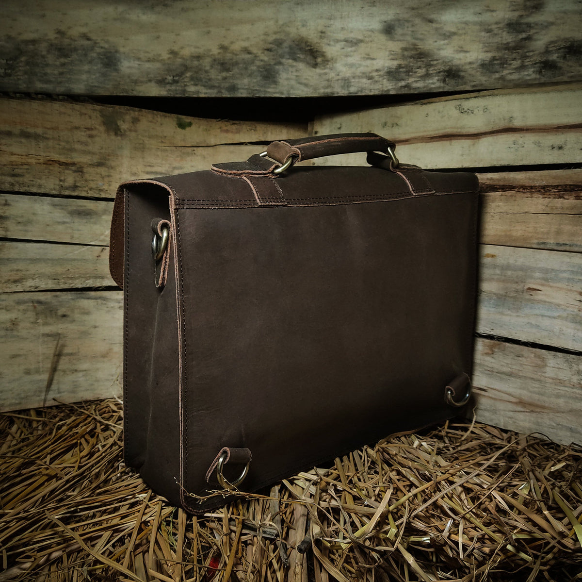 May include: A brown leather briefcase with a top handle and a strap. The briefcase has a flap with a buckle closure and a metal ring on the side. The briefcase is sitting on a bed of straw.