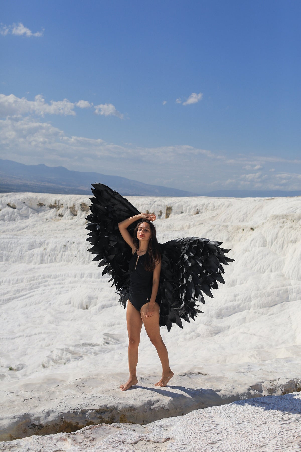 May include: A person wearing a black swimsuit and large black wings stands on a white rocky surface. The background is a blue sky with white clouds.