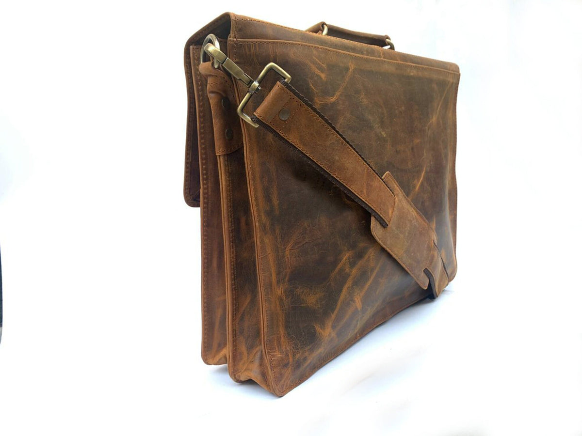 May include: A brown leather briefcase with a shoulder strap and a top handle. The bag has a flap closure and brass-colored hardware. The leather has a distressed appearance, with variations in color and texture.