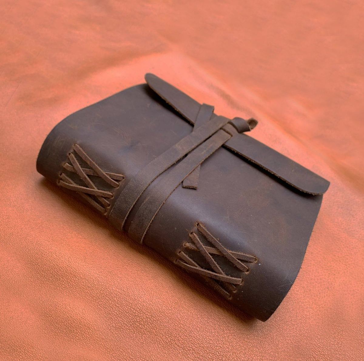 May include: A brown leather journal with a leather strap closure and decorative stitching. The journal is closed and lying on a brown leather surface.