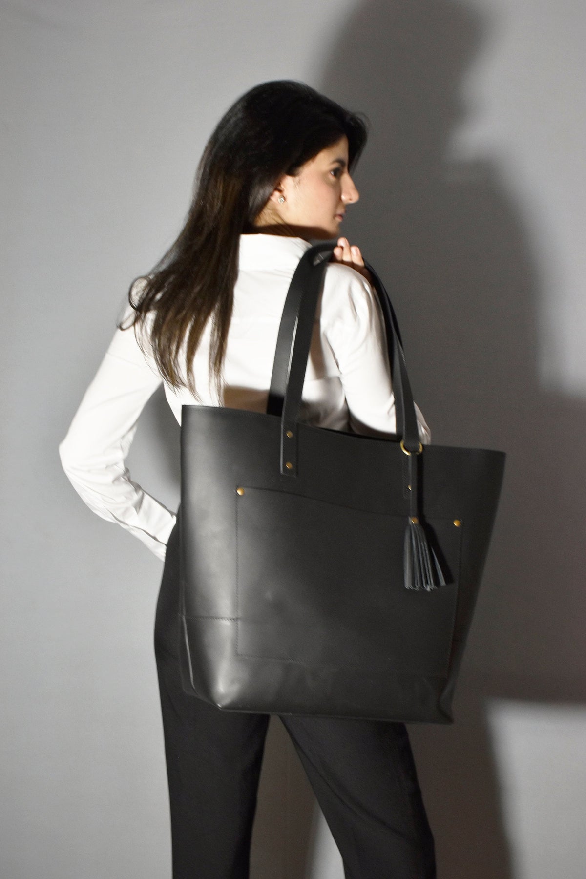 May include: A black leather tote bag with a gold tassel and a front pocket. The bag is being carried by a person wearing a white shirt and black pants.