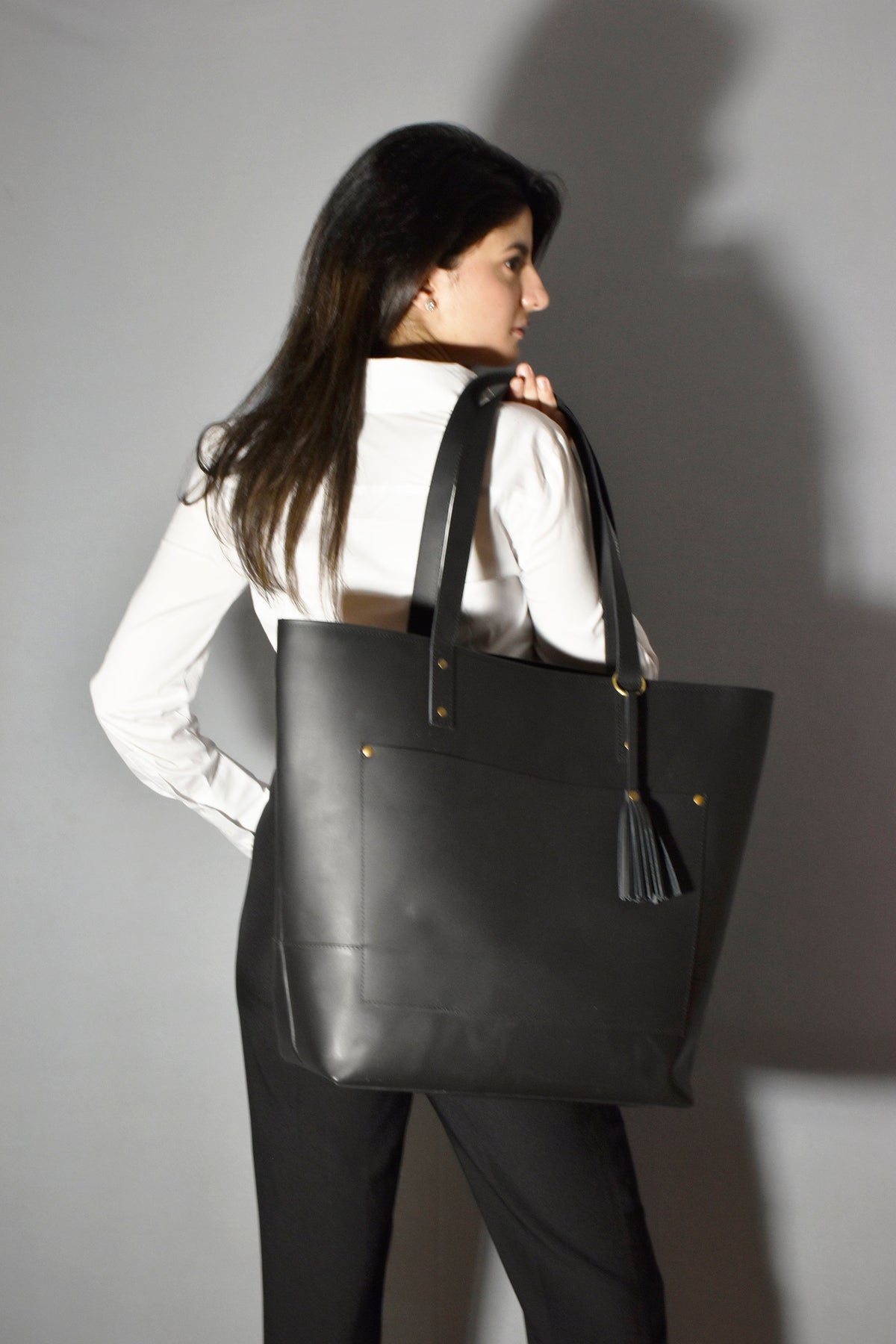 May include: A black leather tote bag with a gold tassel and a front pocket. The bag is being carried by a person wearing a white shirt and black pants.