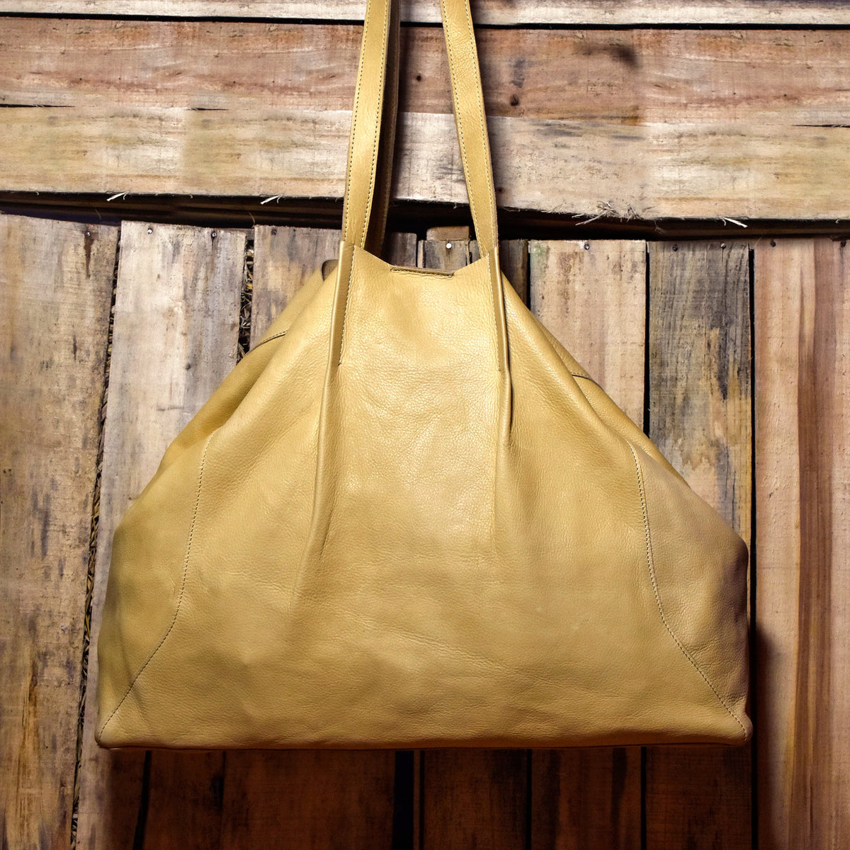 May include: A large, tan leather tote bag with long shoulder straps. The bag has a soft, slouchy design and is hanging against a rustic wooden background. The leather appears to be lightly textured.