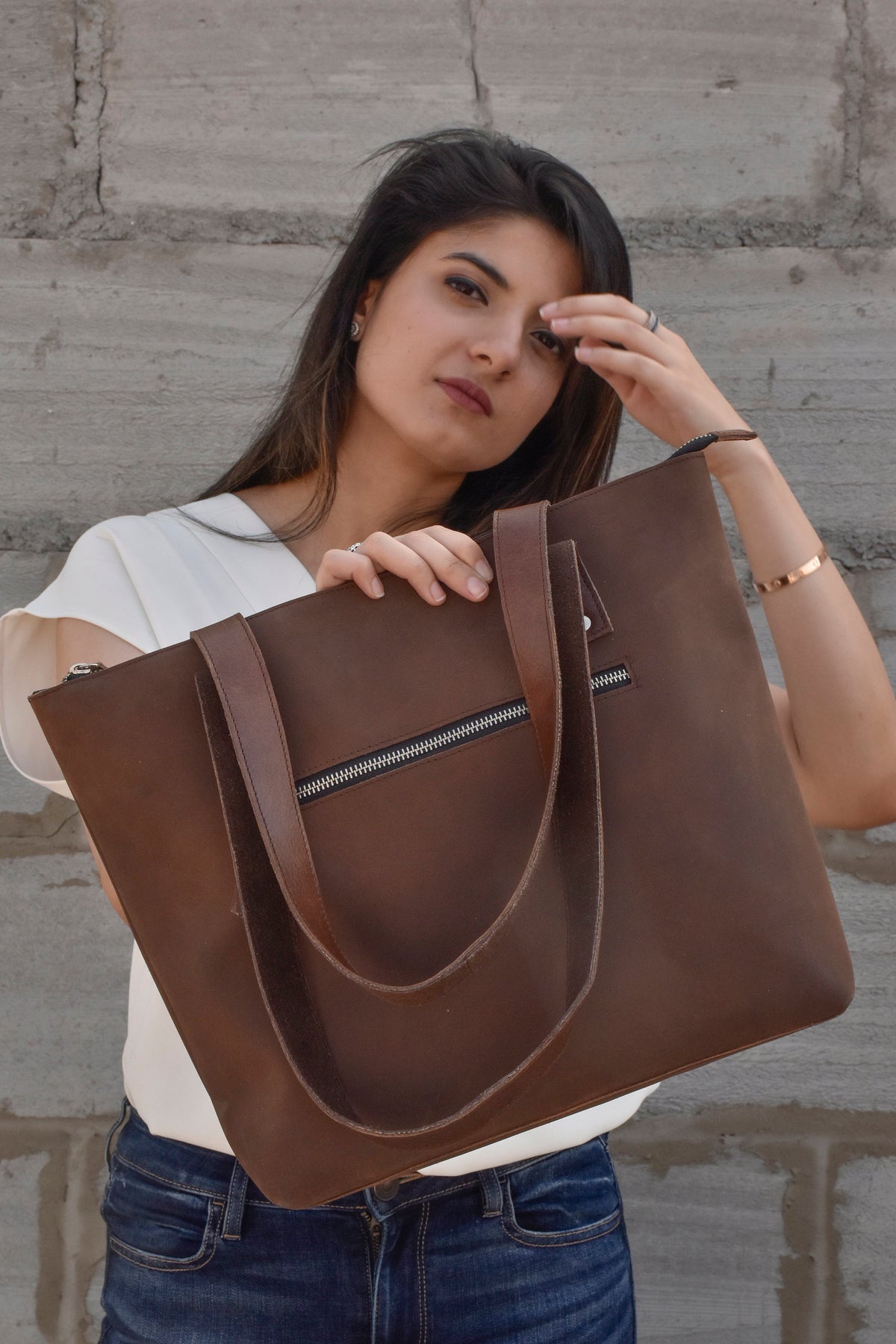 May include: A brown leather tote bag with a zippered pocket and two long straps. The bag is held by a person wearing a white shirt and blue jeans.