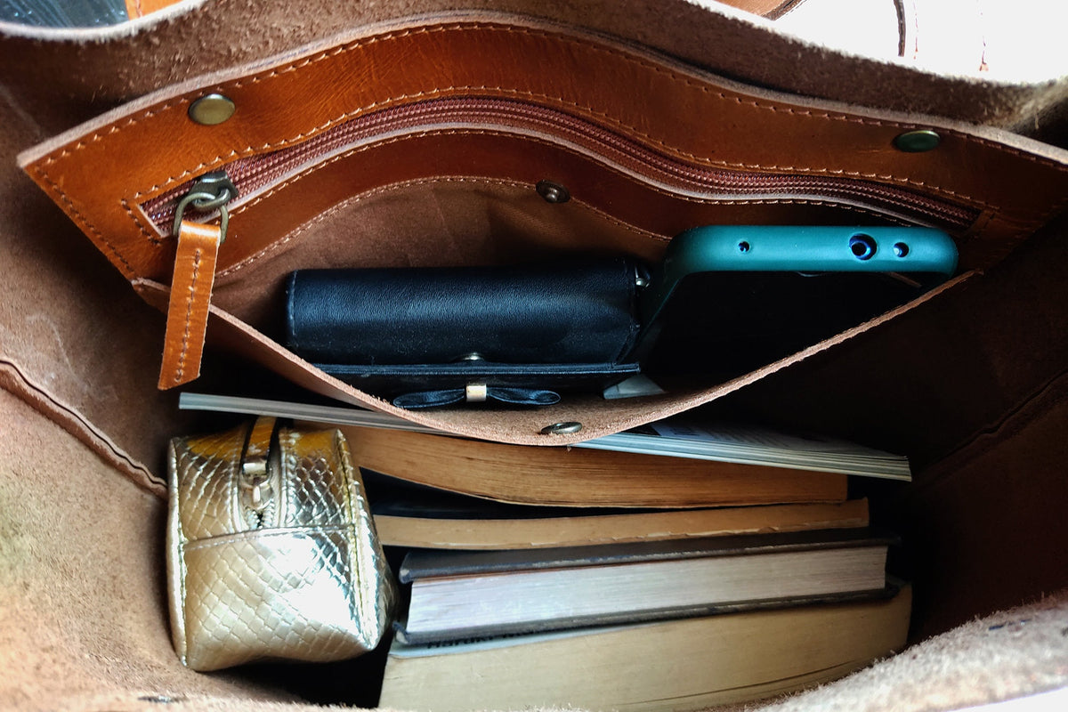 May include: A brown leather tote bag with a zippered pocket and interior compartments. Inside, a teal smartphone, black wallet, gold cosmetic bag, and several books are visible. The bag is open, revealing its contents.