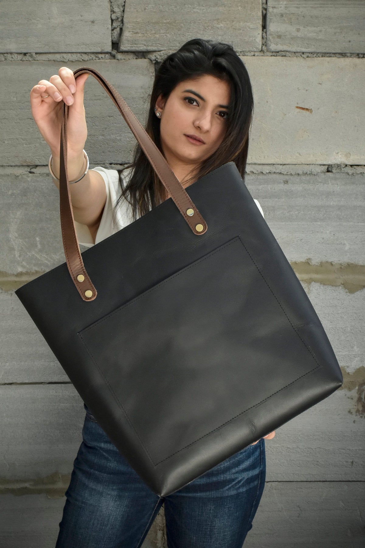 May include: Black leather tote bag with brown leather handles. The tote features a large front pocket and is held up by a person. The handles are attached with brass-colored rivets. The bag is made of a smooth, matte leather.