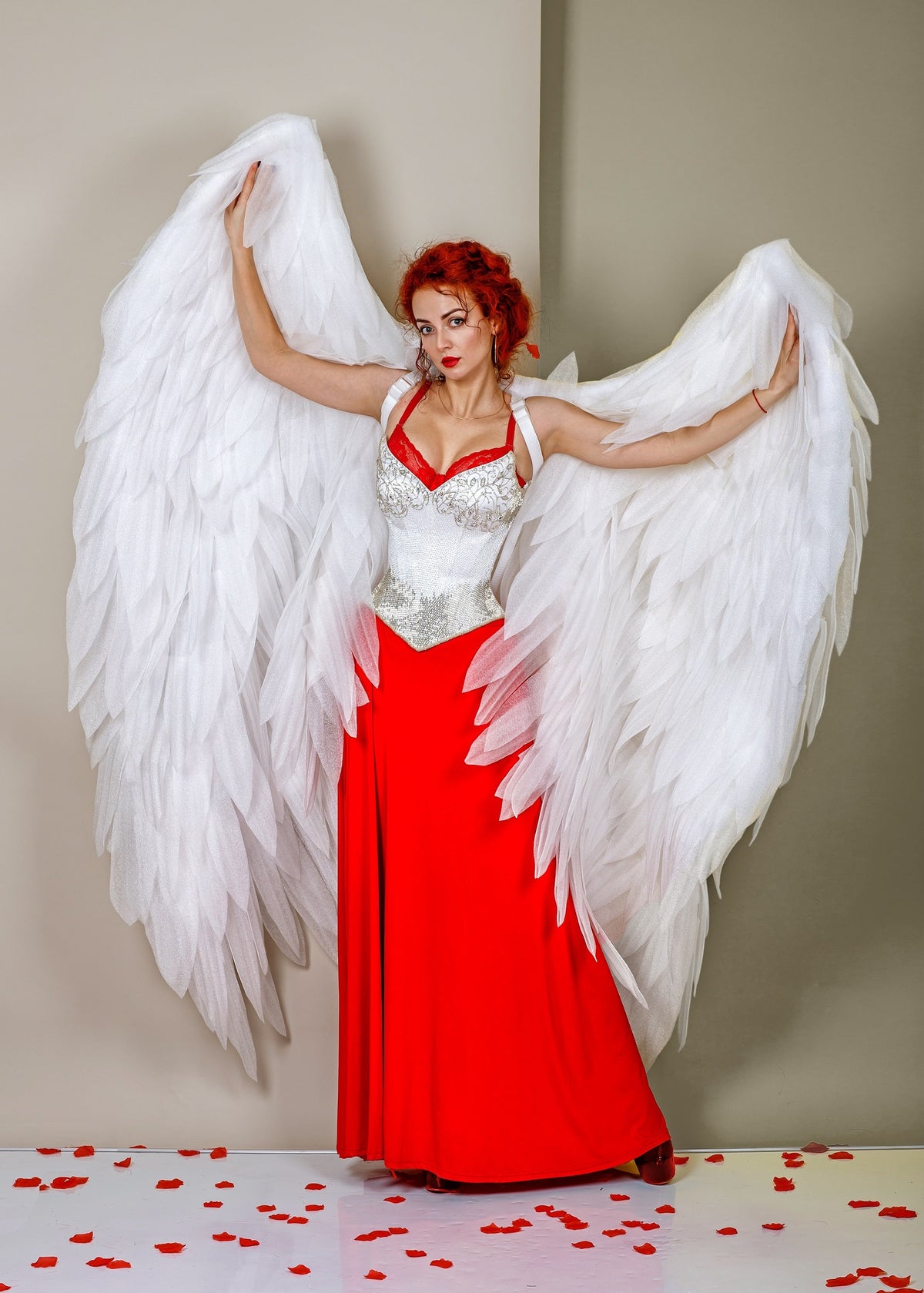 May include: A woman with red hair wearing a white corset and a red dress with large white angel wings. The woman is standing on a white surface with red rose petals scattered around her.