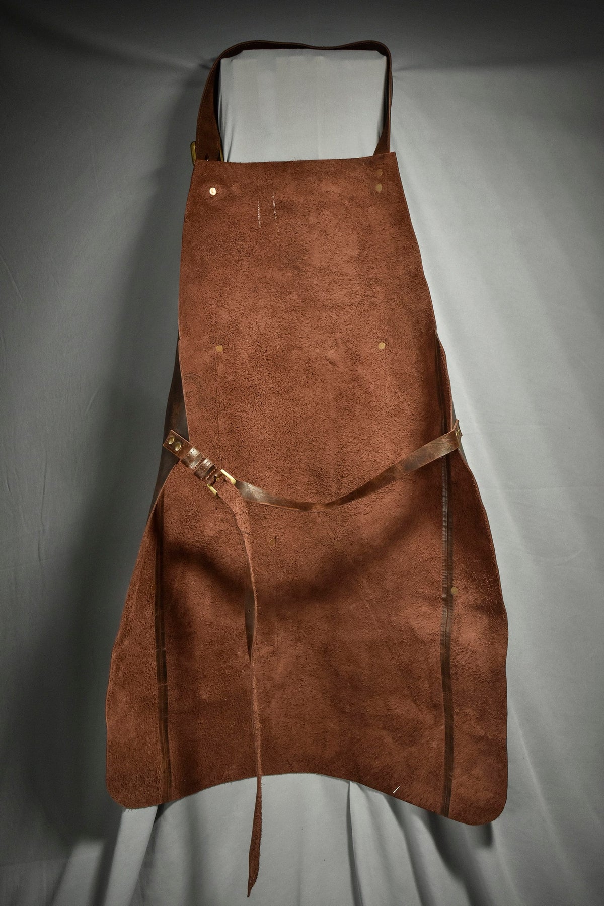 May include: A brown leather apron with adjustable straps and brass buckles. The apron has a suede-like texture and is designed for durability.