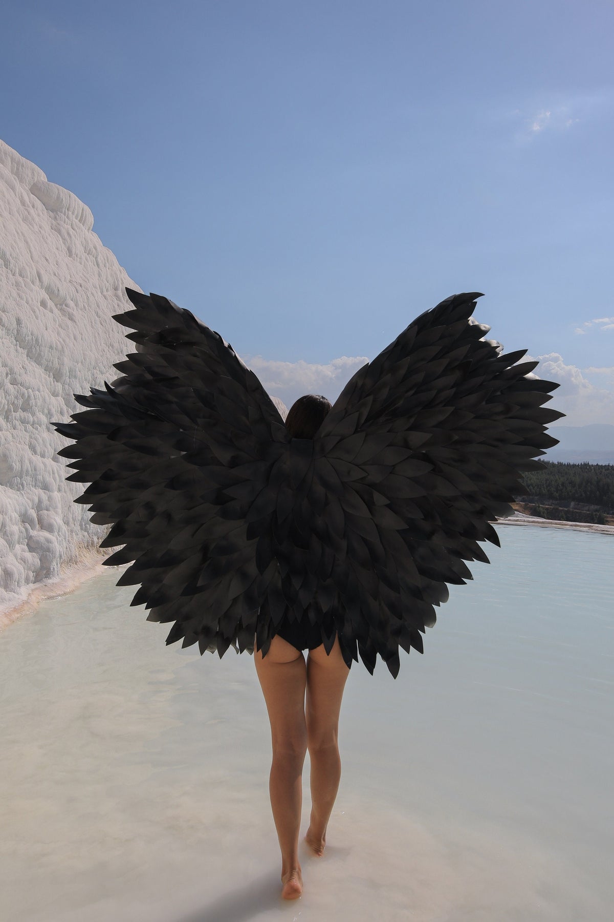 May include: A person wearing a black costume with large black feathered wings walks through a shallow pool of water. The water is a light blue color and the background is a white rock formation.