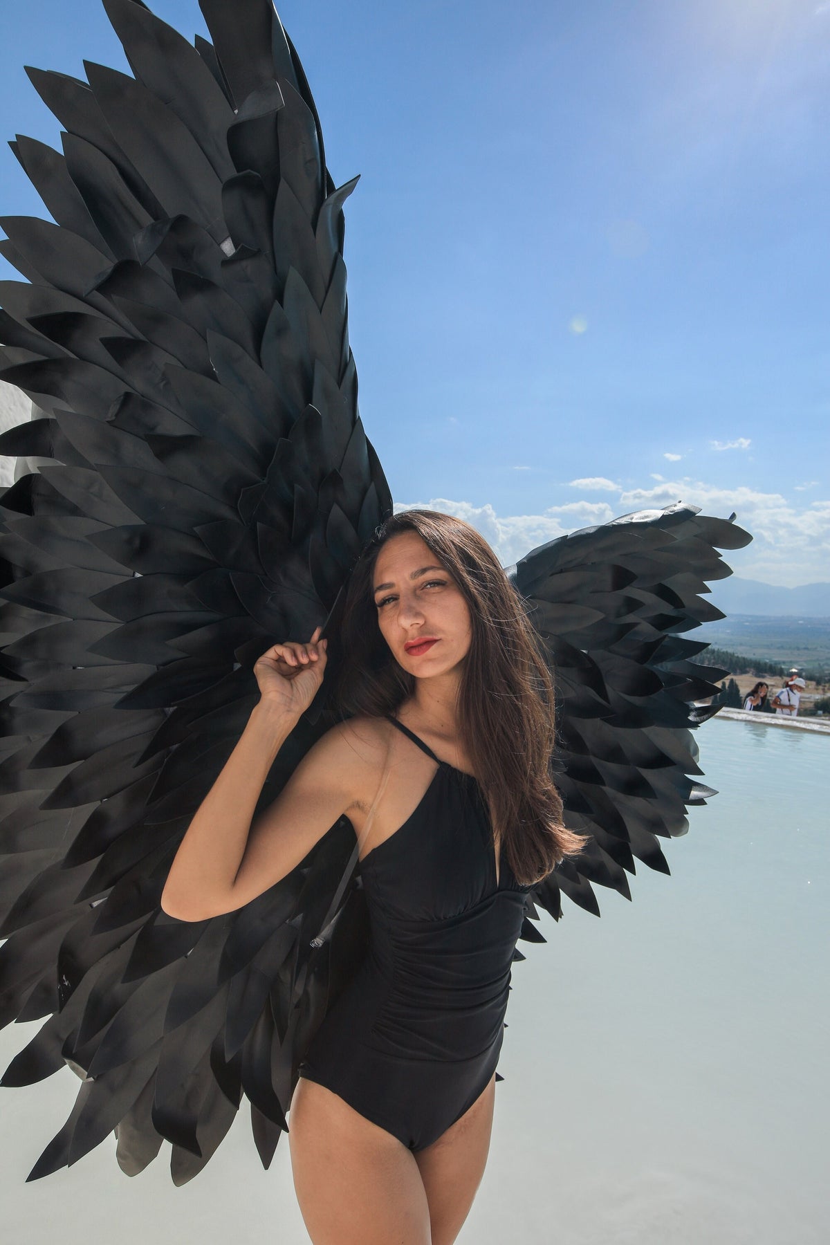 May include: A woman wearing a black swimsuit with large black wings behind her. She is standing in front of a body of water.