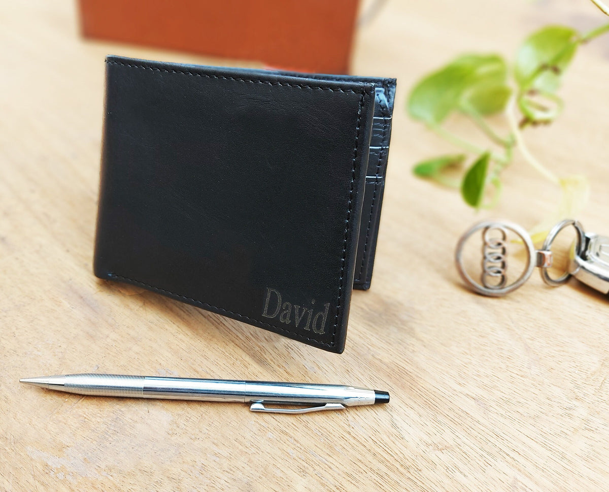 May include: A black leather wallet with the name "David" embossed on the front. The wallet is closed and lying on a wooden surface.