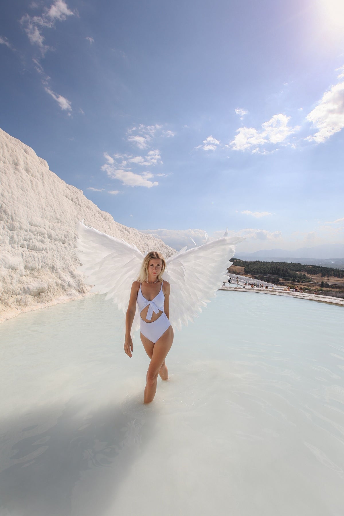 May include: A woman wearing a white swimsuit with a bow in the front stands in a pool of water with white angel wings. The pool is surrounded by white rock formations and a blue sky with white clouds.