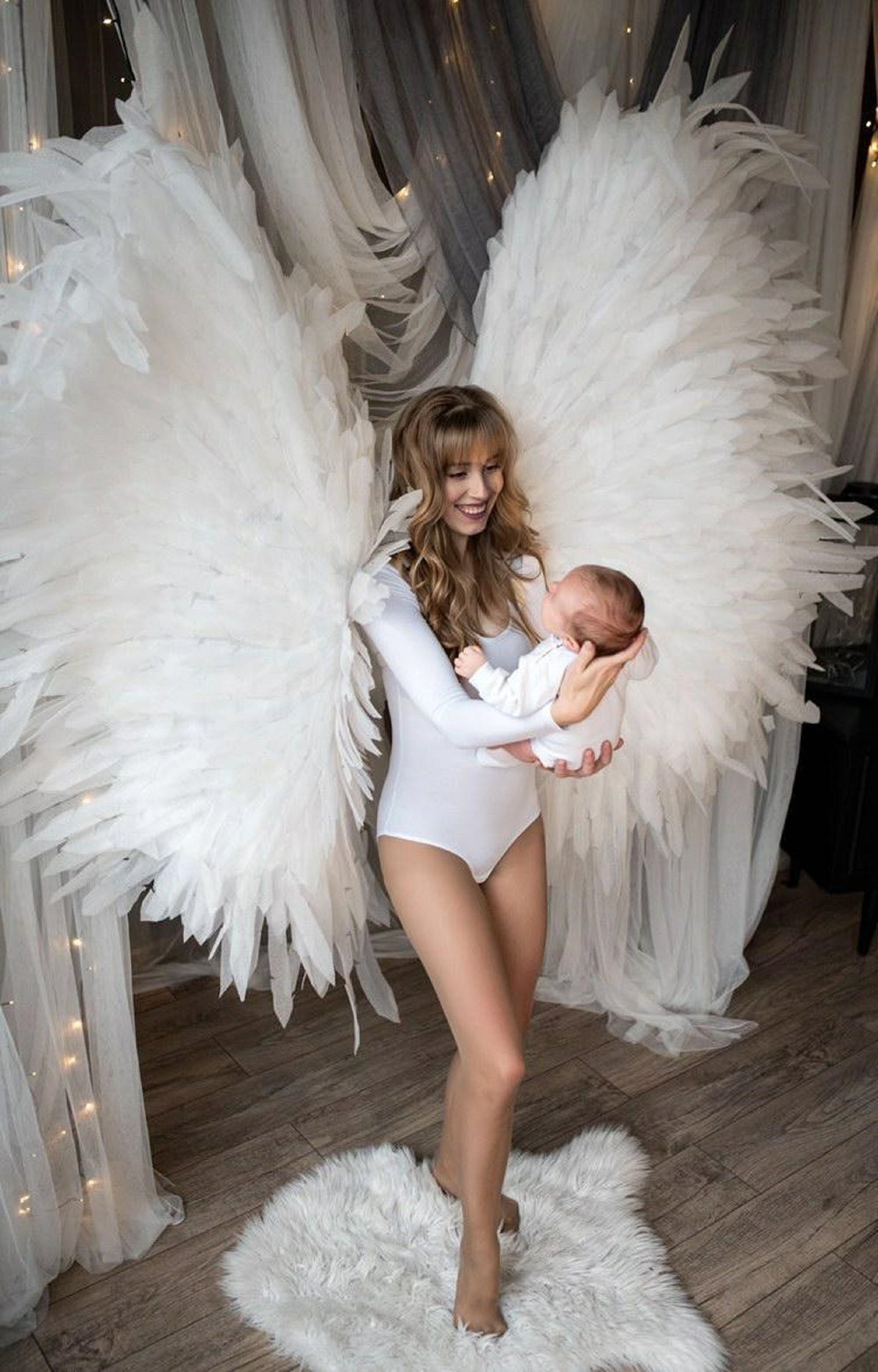 May include: A woman wearing a white bodysuit holds a baby in her arms. She is standing in front of a white backdrop with large white angel wings.