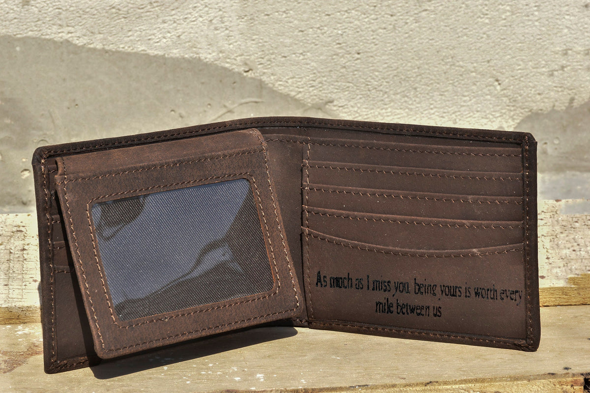 May include: A brown leather wallet with a clear plastic window for a photo ID. The wallet is open and the inside has multiple card slots and a large pocket. The inside of the wallet is engraved with the text "As much as I miss you being yours is worth every mile between us."