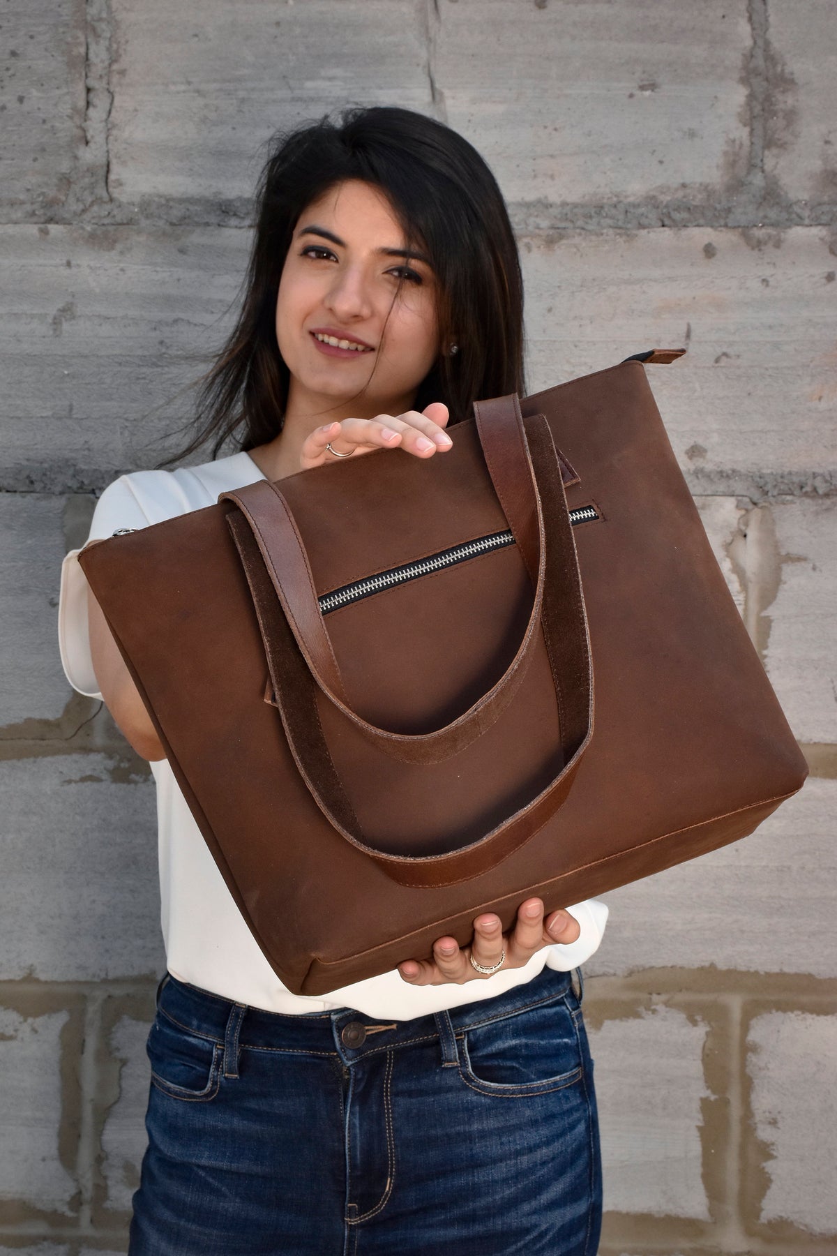 May include: A brown leather tote bag with a zippered pocket and two long straps. The bag is held by a person wearing a white shirt and blue jeans.