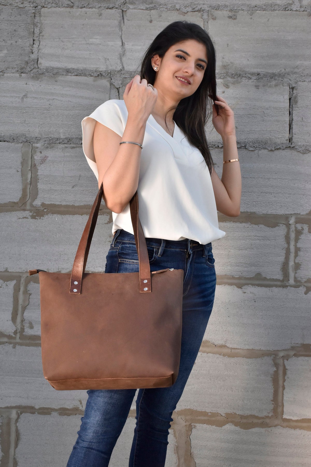 May include: A brown leather tote bag with a zipper closure and two top handles. The bag is being held by a person wearing a white shirt and blue jeans.