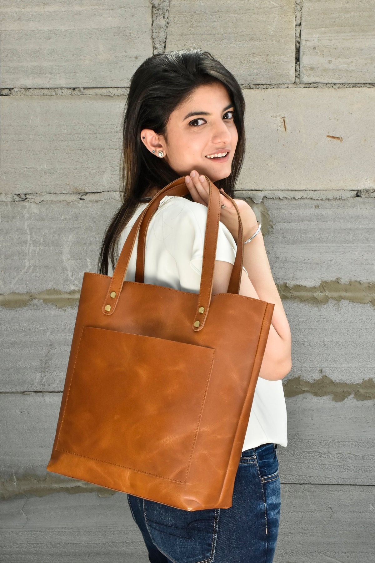May include: A brown leather tote bag with a front pocket and two handles. The bag is held by a person wearing a white shirt and blue jeans. The bag has brass rivets at the handle attachments.
