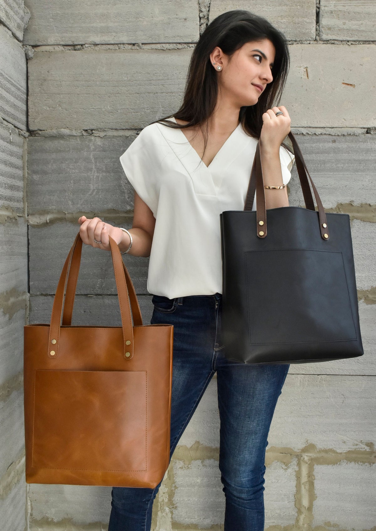 May include: Two leather tote bags, one in a rich brown and the other in black, are held up. Both bags have a rectangular design with a front pocket and brown leather handles. The bags are accented with gold-colored rivets.