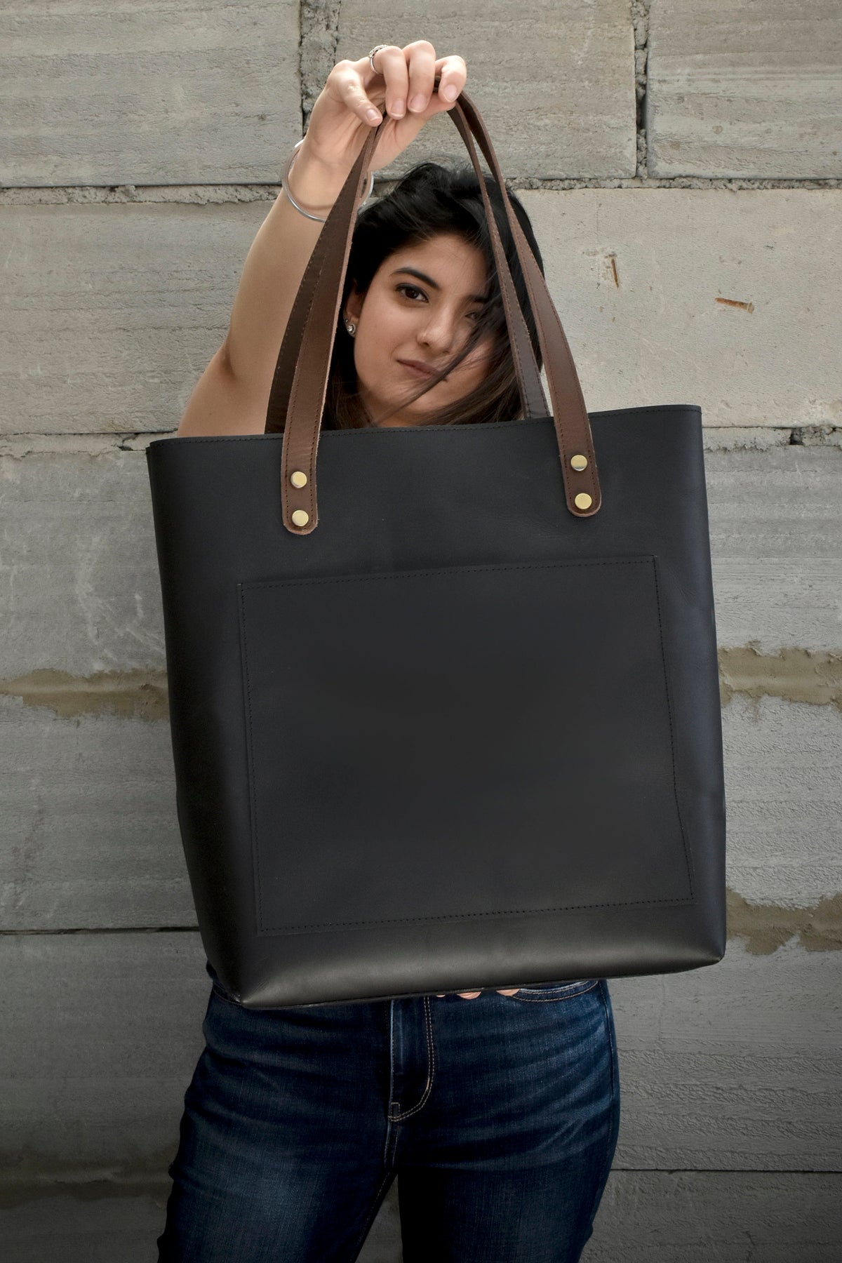 May include: A black leather tote bag with brown leather handles and gold-colored rivets. The bag has a large front pocket and is held up against a gray brick wall. The handles are attached with gold rivets.