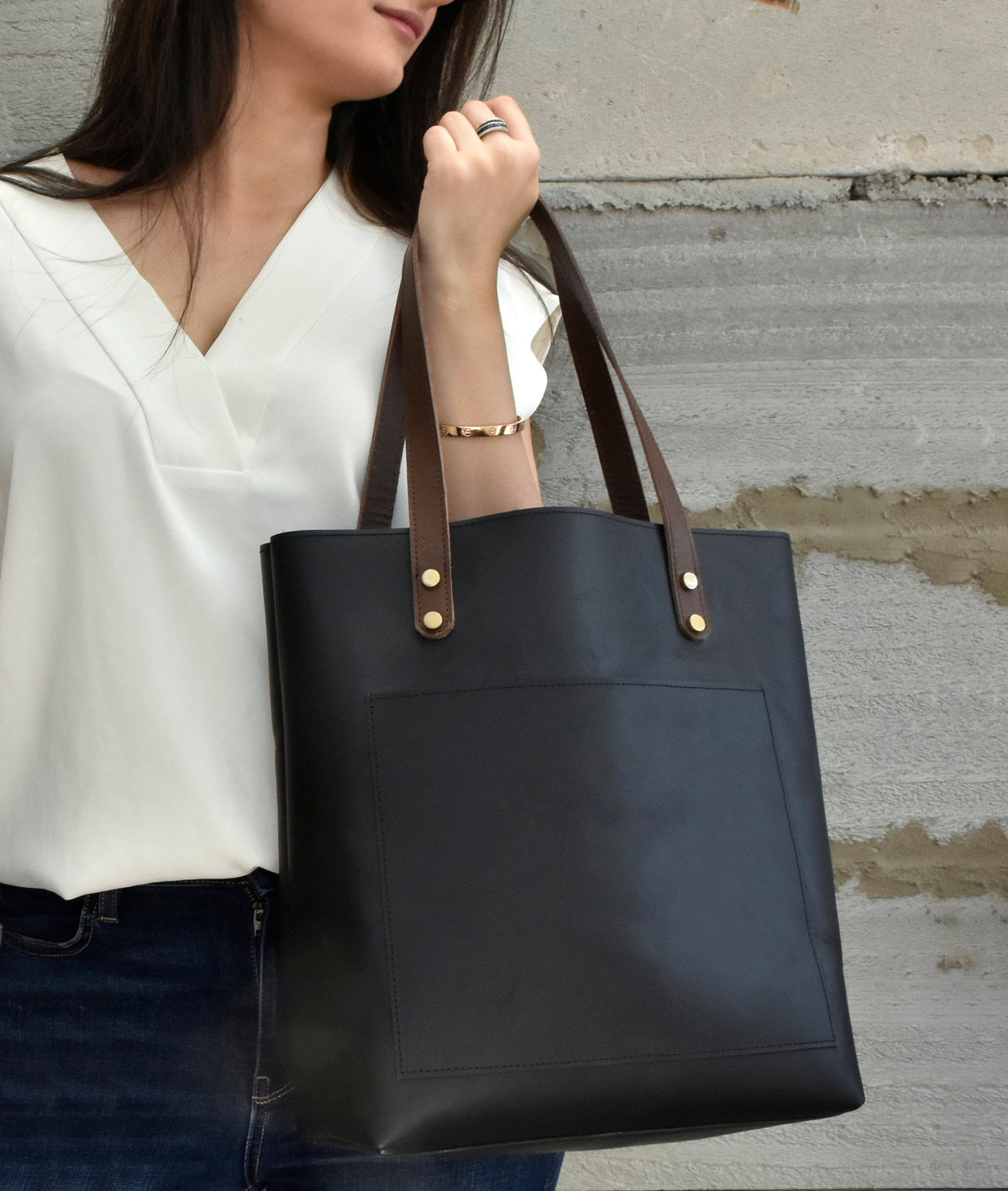 May include: Black leather tote bag with brown leather handles and gold-colored rivets. The bag has a large front pocket and is being held by a person wearing a white top and blue jeans. The bag is a classic and versatile accessory.