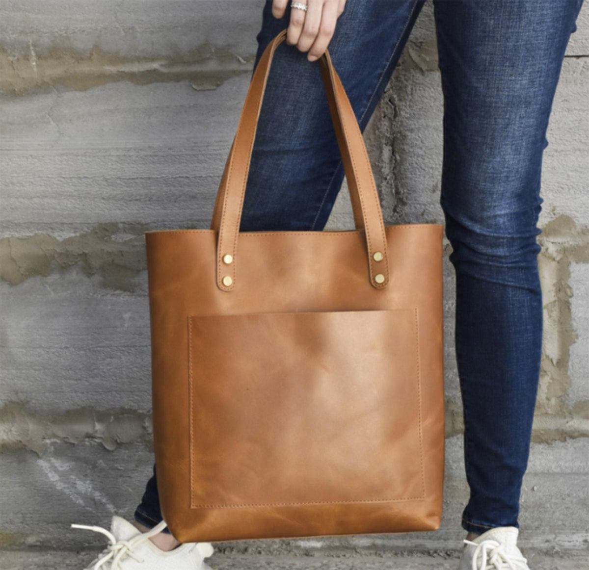 May include: A tan leather tote bag with a front pocket and two handles. The bag is held by a person wearing blue jeans and white sneakers. The bag has brass rivets at the top of the handles.