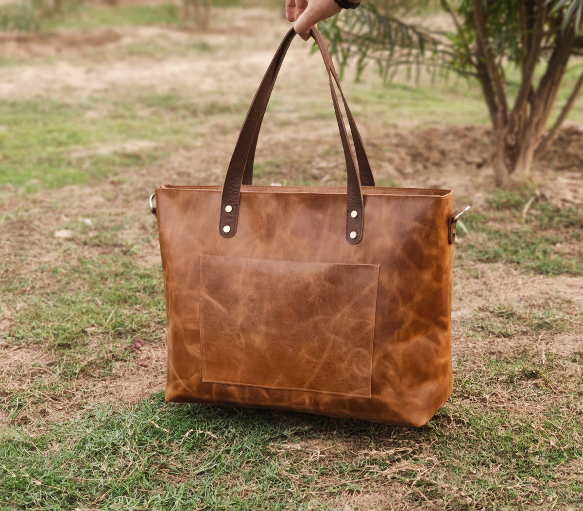 May include: A brown leather tote bag with a front pocket and brown leather handles. The bag is sitting on a bed of green grass.