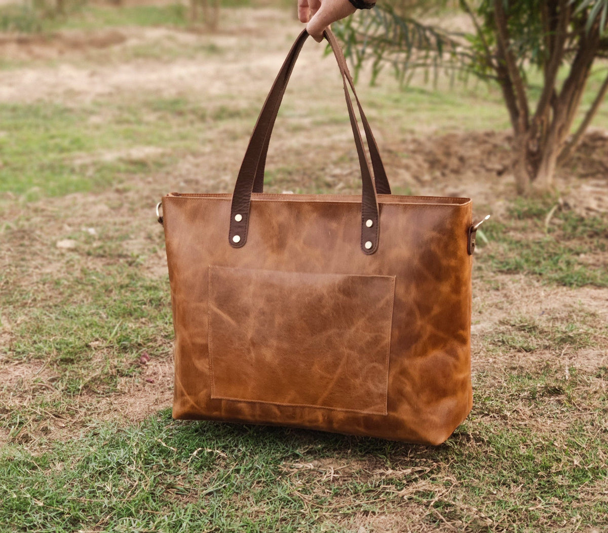 May include: A brown leather tote bag with a large front pocket and dark brown handles. The bag has a rectangular shape and is held up by a person's hand. The bag is sitting on a green grassy surface.
