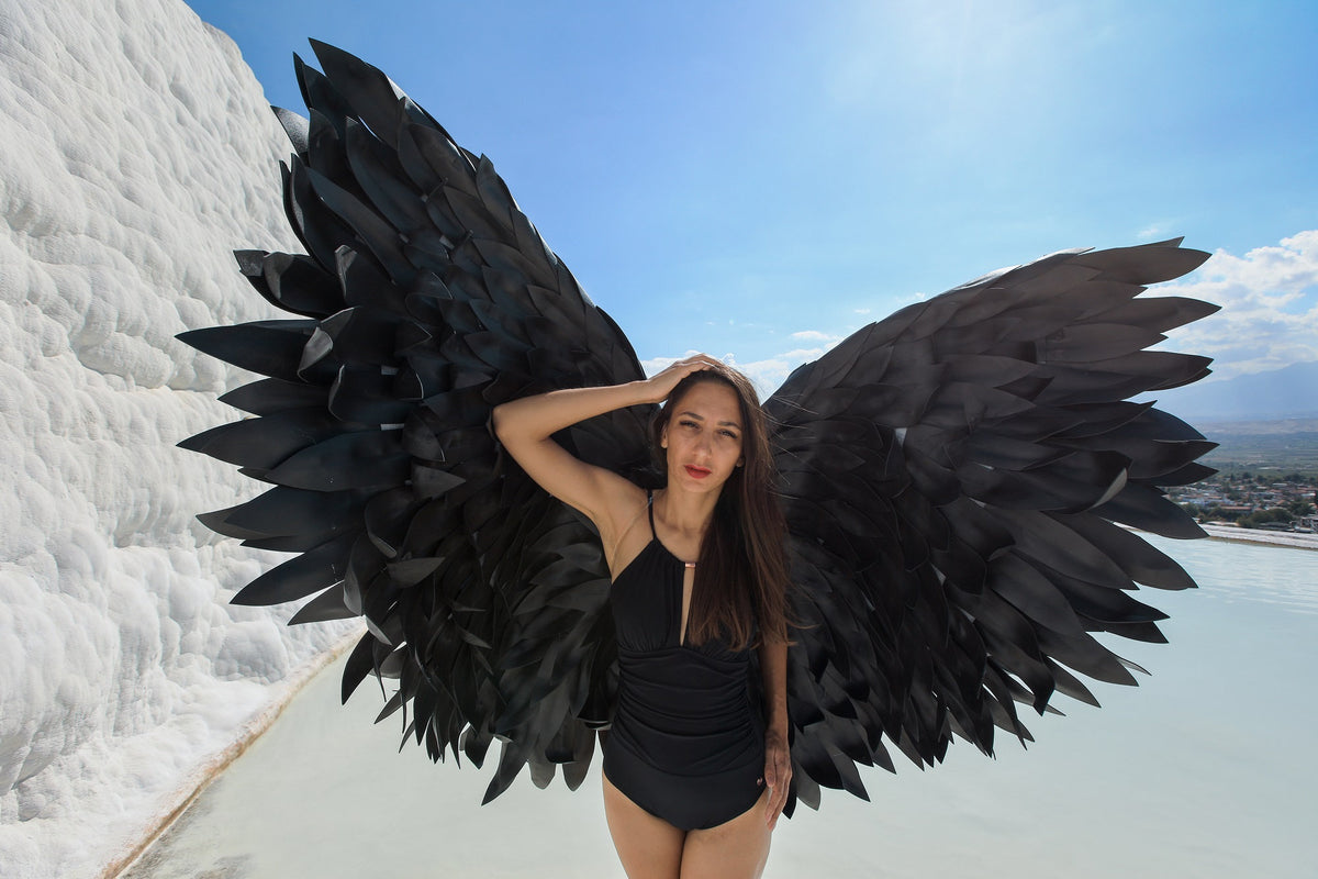 May include: A woman wearing a black swimsuit with large black angel wings. The wings are made of feathers and are spread out behind her. She is standing in front of a white background.
