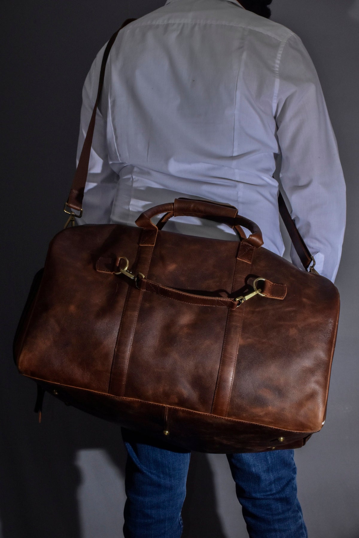 May include: A brown leather duffel bag with a shoulder strap and top handles. The bag has brass-colored hardware and is carried over the shoulder. The leather has a distressed appearance. The person is wearing a white shirt and blue jeans.