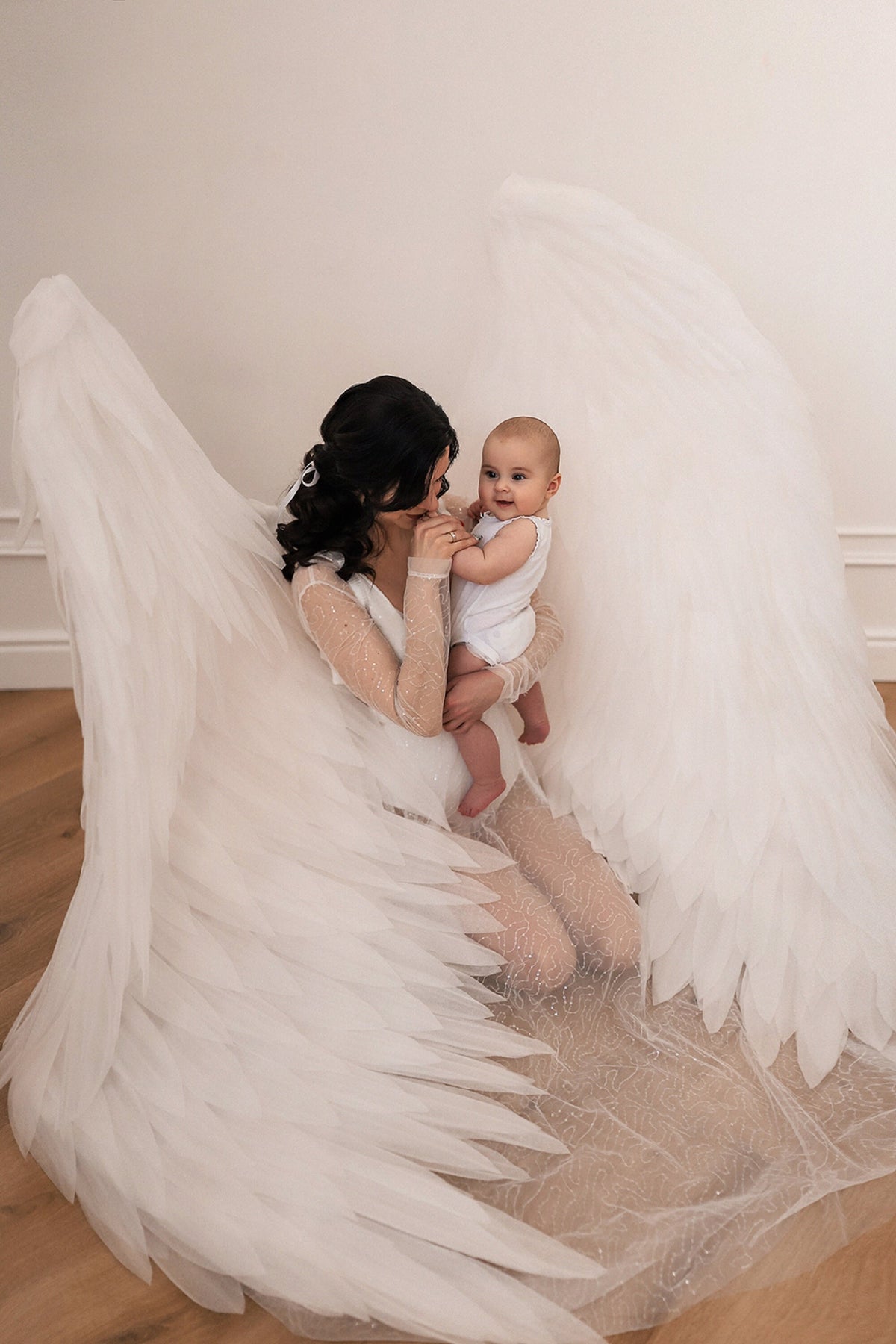 May include: A woman wearing a white dress with angel wings holds a baby in her arms. The baby is wearing a white onesie and looking at the camera. The woman is looking at the baby and smiling.