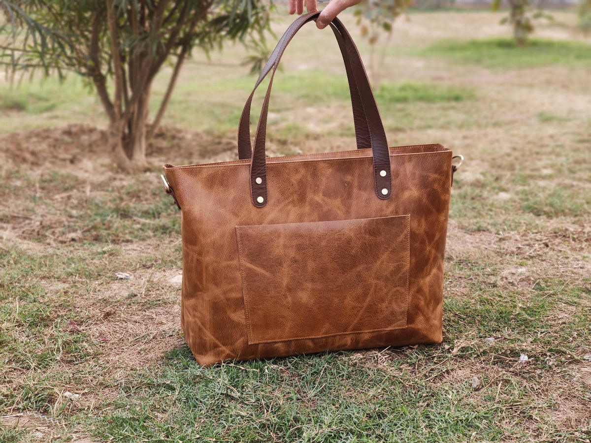 May include: A brown leather tote bag with a front pocket and dark brown handles. The bag has a rectangular shape and is held by a person. The bag is sitting on a grassy surface.