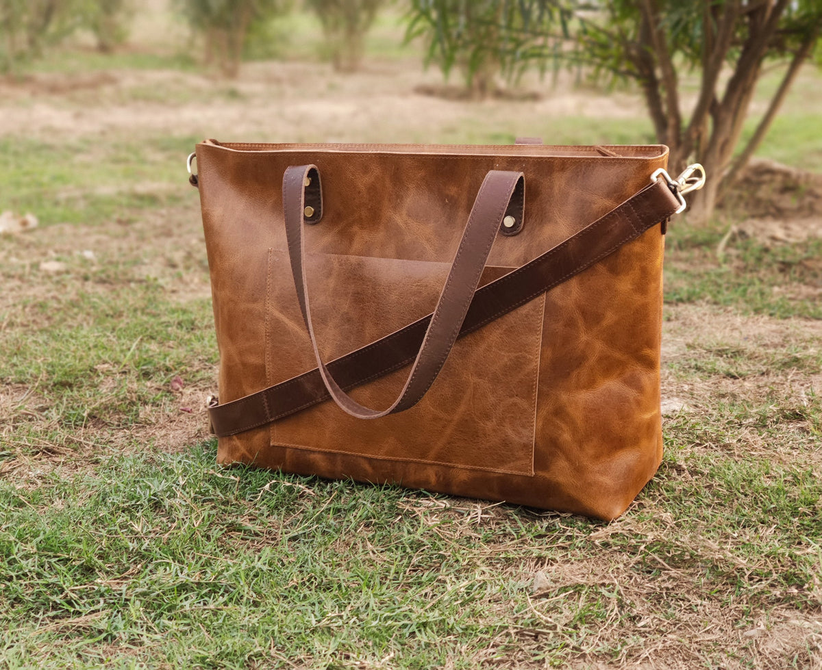 May include: A brown leather tote bag with a front pocket and two brown leather handles. The bag also has a detachable brown shoulder strap with gold-colored hardware. The bag is sitting on green grass.
