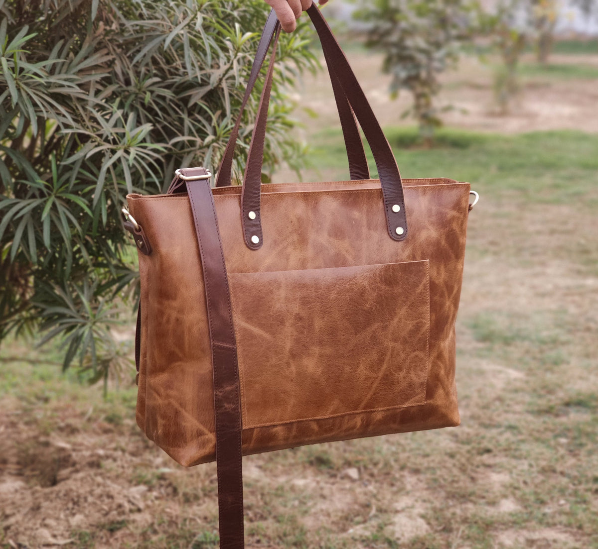 May include: A brown leather tote bag with a front pocket and a detachable shoulder strap. The bag has two top handles and is held up by a person. The leather has a distressed look.