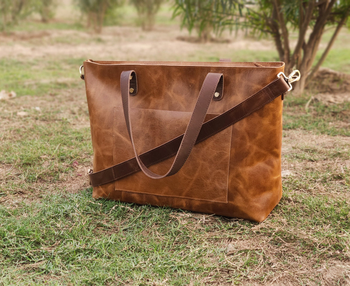 May include: A brown leather tote bag with a front pocket and a detachable shoulder strap. The bag has two top handles and gold-colored hardware. The bag is sitting on green grass.