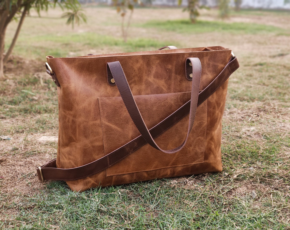 May include: A brown leather tote bag with a front pocket and a detachable shoulder strap. The bag has two top handles and a brown leather strap that goes across the body. The bag is sitting on green grass.