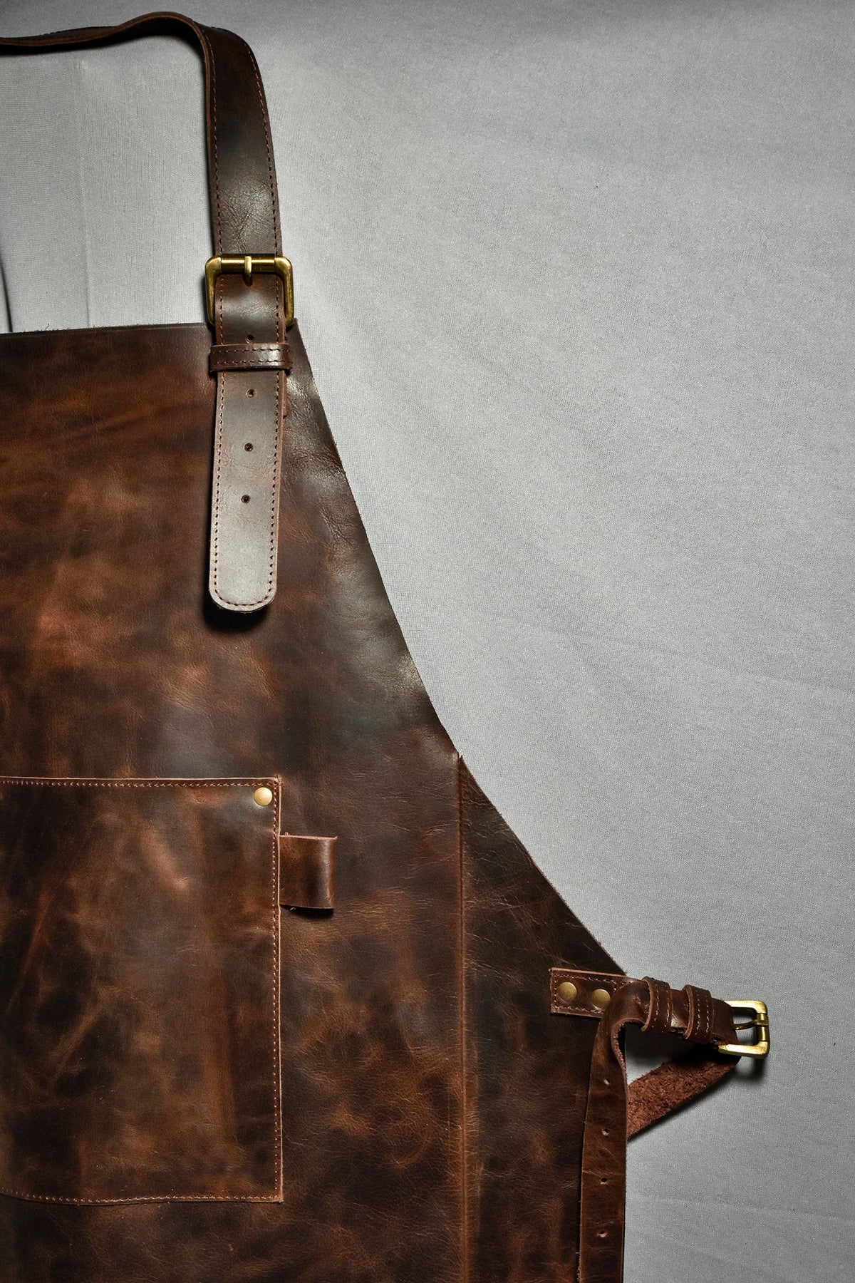 May include: A brown leather apron with a large pocket and adjustable straps. The apron has brass buckles and rivets.