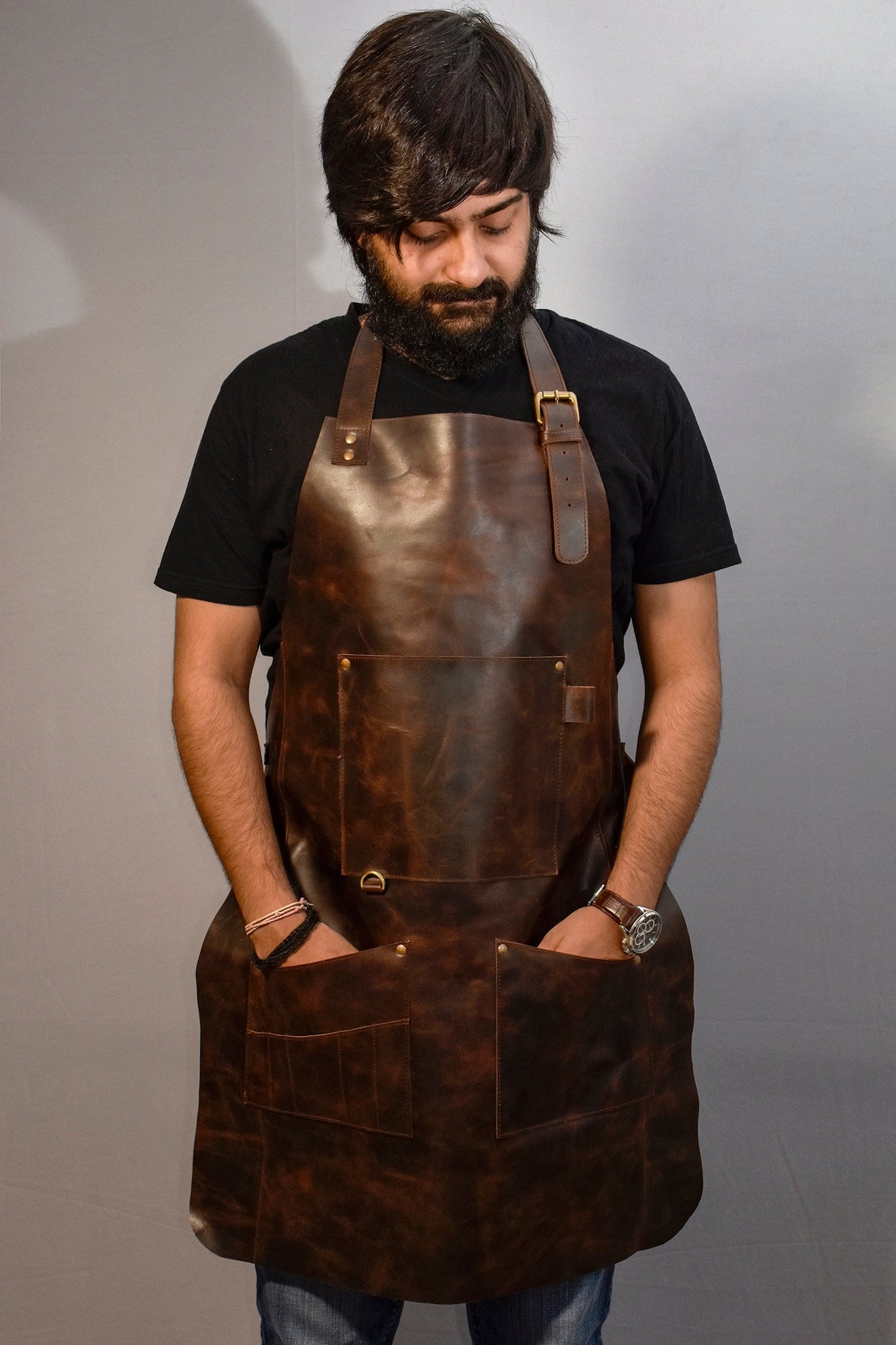 May include: A brown leather apron with multiple pockets and a brass buckle. The apron is worn by a person with a beard and a black t-shirt.