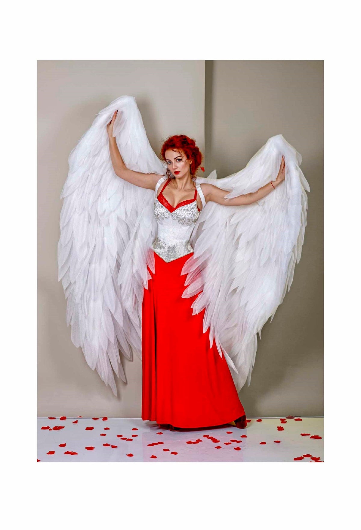 May include: A woman with red hair wearing a white corset and a red skirt with large white angel wings. The background is a light beige wall and the floor is covered in red rose petals.