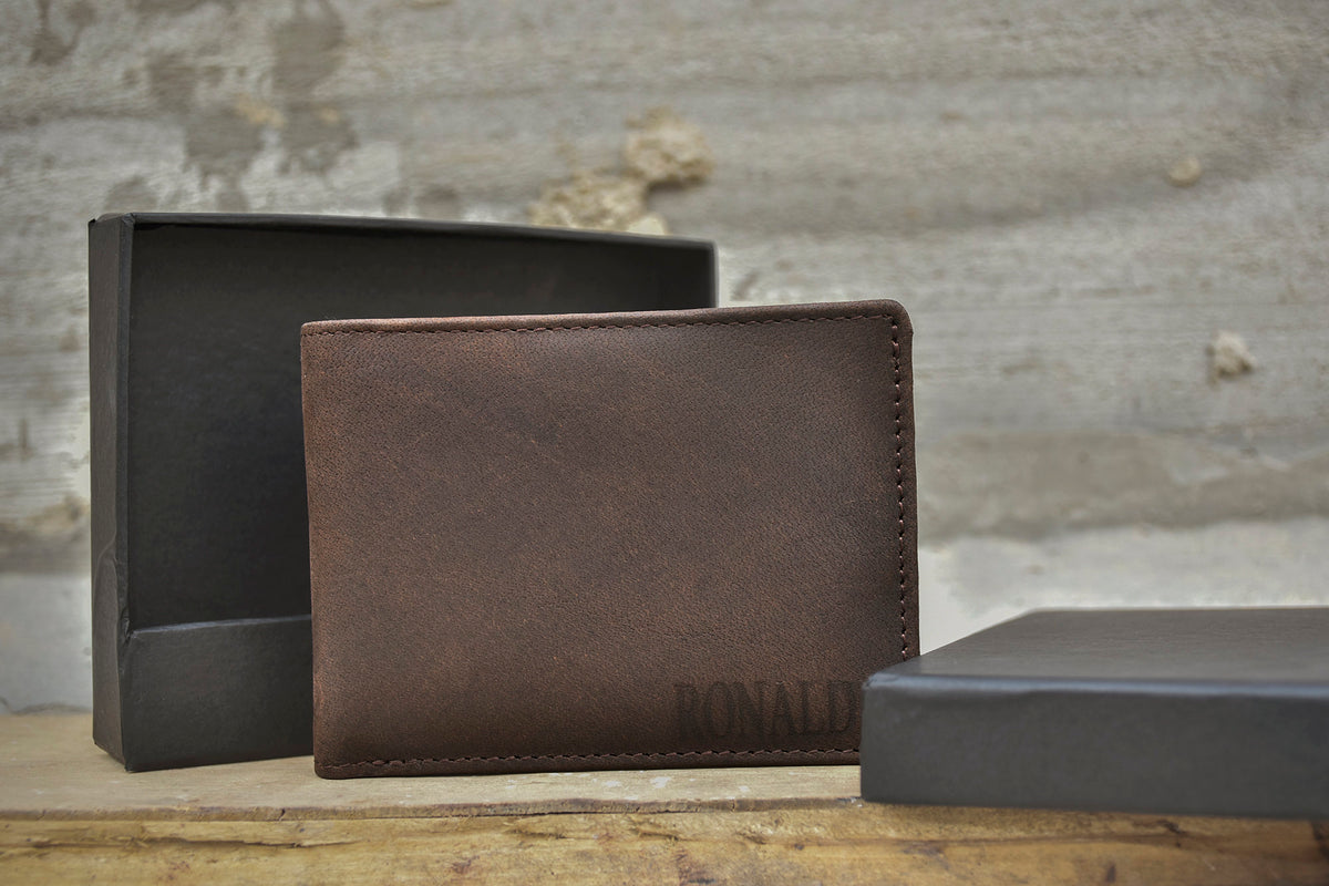 May include: A brown leather wallet with the name "RONALD" embossed on the front. The wallet is sitting on a wooden surface next to two black boxes.