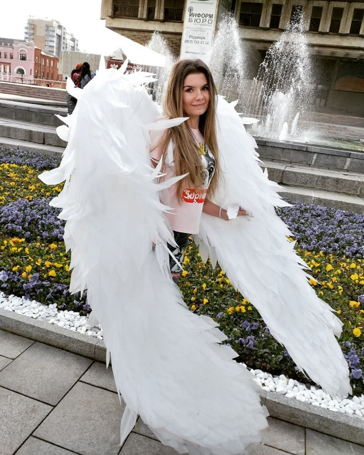 May include: A woman wearing a pink shirt with a white graphic print and white angel wings. The wings are large and feathery, and the woman is standing in front of a fountain.