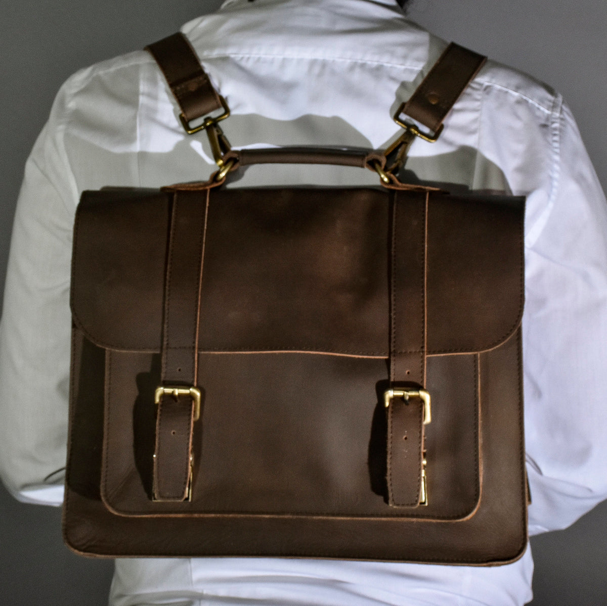 May include: A brown leather briefcase with adjustable shoulder straps and a top handle. The bag features two front straps with gold-colored buckles. The briefcase is worn as a backpack.