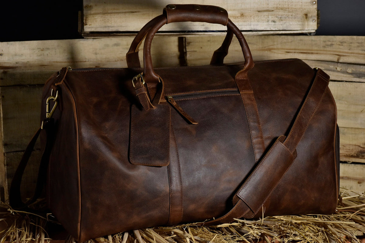 May include: A brown leather duffel bag with a top handle and a shoulder strap. The bag has a zippered pocket on the front and a small leather tag.