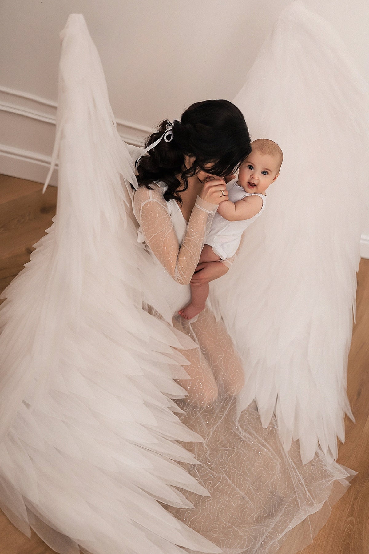 May include: A woman wearing a white gown with angel wings holds a baby in her arms. The woman is looking down at the baby, who is looking up at the camera. The background is a white wall.