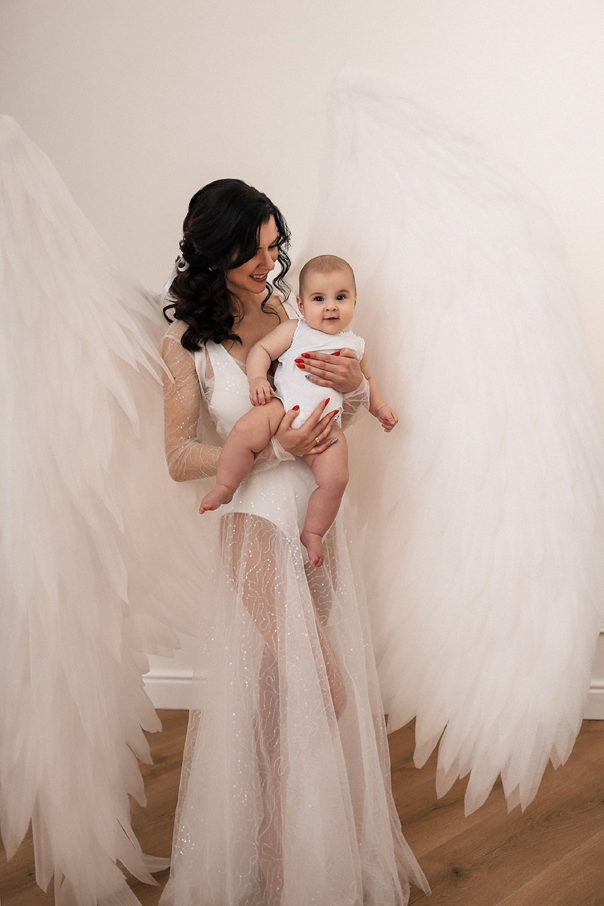 May include: A woman in a white, sparkly dress holds a baby in her arms. The woman is wearing a white angel wing costume. The baby is wearing a white onesie.