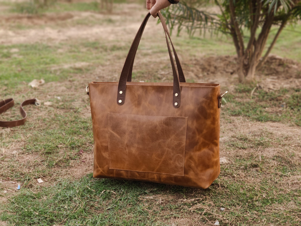 May include: A brown leather tote bag with a rectangular front pocket and two top handles. The bag has a shoulder strap and is held up by a person's hand. The bag is sitting on a grassy surface.